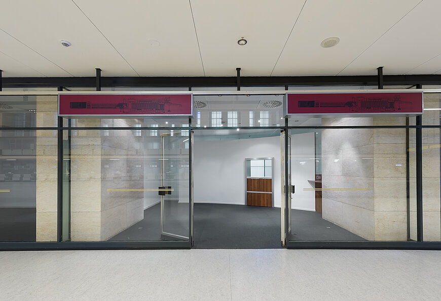 Blick in einer der Glasbüros in der Haupthalle des Flughafen Tempelhof. Die Glastür ist geöffnet. Über der Glasfenster hängen 2 rote Schilder