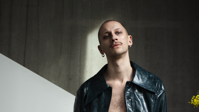 Porträt von DJ Darwin Stapel in einer dunklen, industriellen Umgebung. Er trägt eine offene, glänzende schwarze Lederjacke und steht vor einer Betonwand mit einem weißen geometrischen Paneel. Rechts im Bild steht ein Strauß leuchtend gelber Blumen, der einen starken Farbkontrast zur Szene bildet.