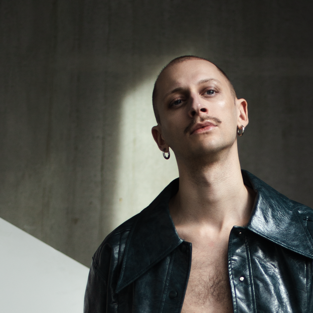 Porträt von DJ Darwin Stapel in einer dunklen, industriellen Umgebung. Er trägt eine offene, glänzende schwarze Lederjacke und steht vor einer Betonwand mit einem weißen geometrischen Paneel. Rechts im Bild steht ein Strauß leuchtend gelber Blumen, der einen starken Farbkontrast zur Szene bildet.