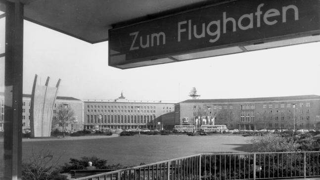 Ein schwarz‑weißes historisches Foto des Berliner Flughafens Tempelhof: Im Vordergrund steht ein Wegweiser mit der Aufschrift „Zum Flughafen“. Dahinter erstreckt sich das weitläufige Vorfeld mit parkenden Bussen und Autos. Zentral im Hintergrund ragt das Luftbrücken‑Denkmal mit seinen drei gebogenen Säulen auf, flankiert von den langgezogenen Gebäudefassaden des Flughafens.