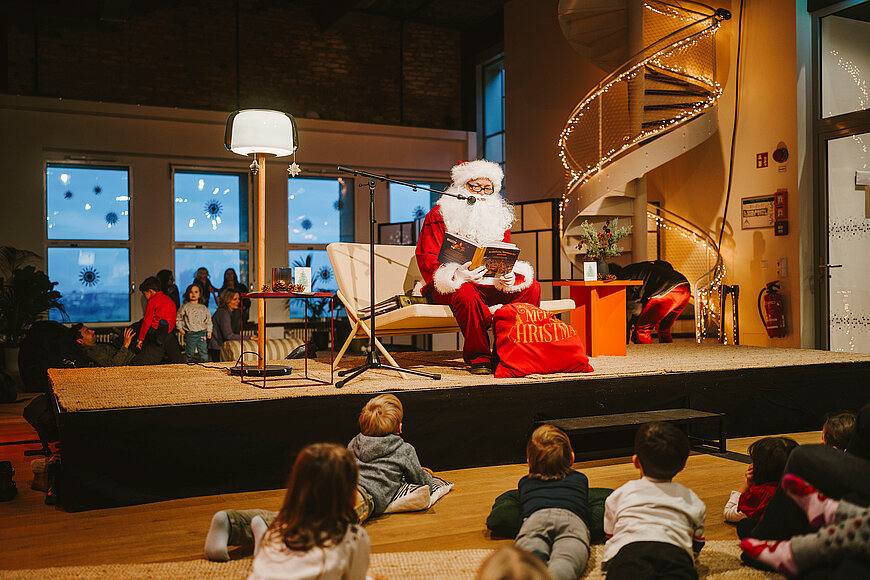 Ein Weihnachtsmann sitzt im THF TOWER auf einem Sofa und liest aus einem Buch. Vor ihm liegen Kinder auf dem Boden uns hören ihm zu. 