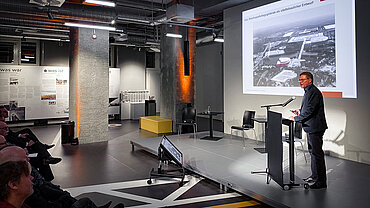 Mann am Rednerpult vor Leinwand mit historischer Aufnahme