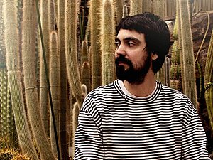 Ein Mann sitzt vor einem Hintergund mit holzvertäfelter Wand und vielen Kakteen.