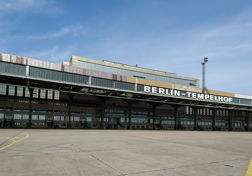 Außenansicht des Flughafengebäudes Berlin-Tempelhof an einem sonnigen Tag. Im Zentrum dominiert die charakteristische, lange, überdachte Vorfelderweiterung. Auf dem oberen Teil der Fassade ist in großen, braunen und weißen Buchstaben der Schriftzug "BERLIN-TEMPELHOF" zu sehen, zusammen mit der Angabe der Höhe ("164 FEET - 50 M FEET ELEVATION").