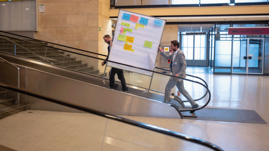 Zwei Menschen tragen auf einer Rolltreppe in der Haupthalle am Flughafen Tempelhof eine Stellwand