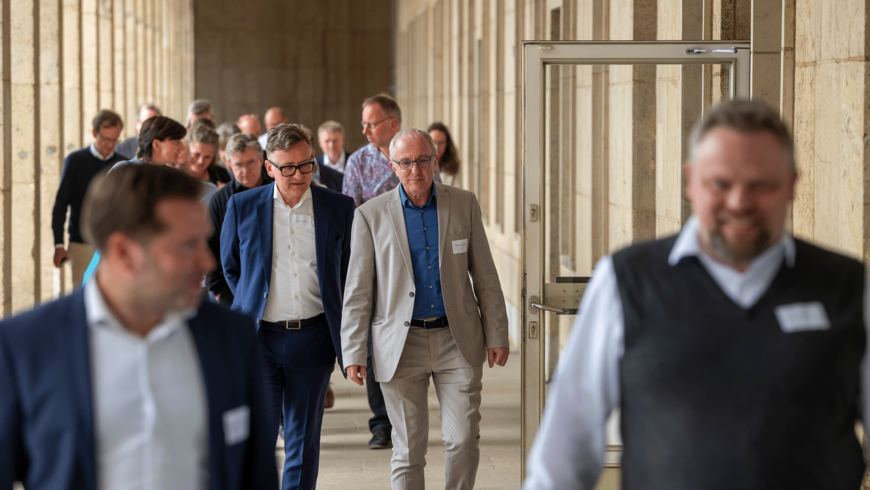 Eine Gruppe von Personen in Business-Kleidung geht gemeinsam durch einen hellen, säulengesäumten Gang am Flughafen Tempelhof