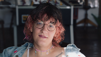 Porträt von Kuku Schrapnell in einem Innenraum. Die Person trägt eine runde Brille, eine helle Jeansjacke und eine rosa Perlenkette mit kleinen Anhängern. Das Haar ist dunkel am Ansatz und pink in den Spitzen. Ein Nasenring und ein sichtbares Brust‑Tattoo ergänzen den Look. Der Hintergrund ist unscharf und zeigt ein kreatives Arbeitsumfeld.