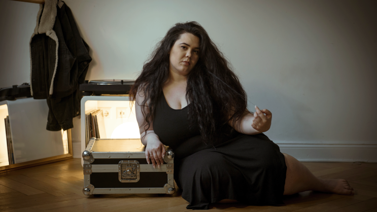 DJ TRU:L sitzt auf einem Holzboden neben einem silbernen Equipment-Case, trägt ein schwarzes ärmelloses Kleid und blickt leicht zur Seite; im Hintergrund stehen ein Spiegel, ein Kleiderständer und ein Regal mit Schallplatten.