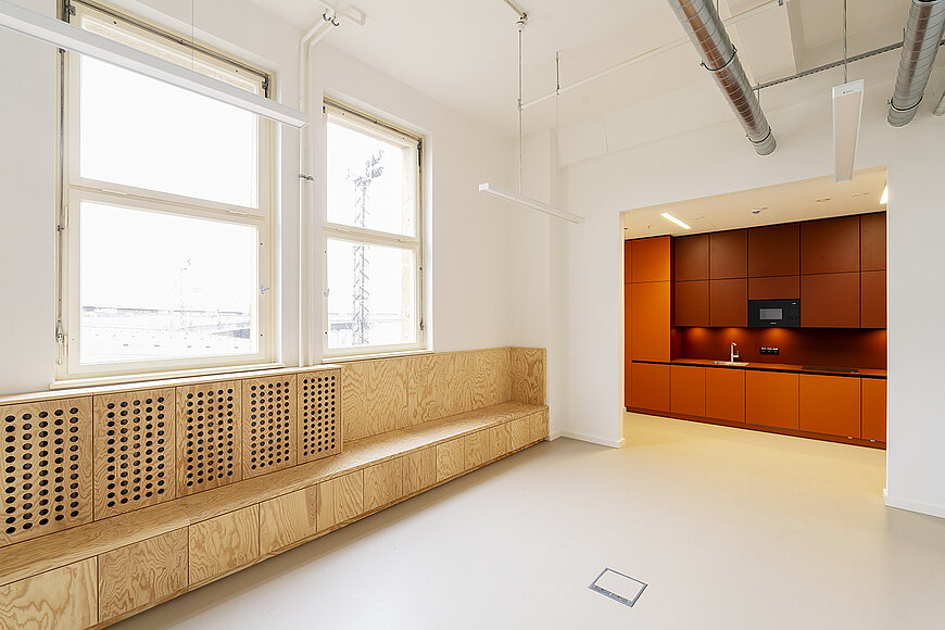 Innenansicht eines hellen, sanierten Büros am Flughafen Tempelhof (Kopfbau West). Der Raum hat einen hellen Boden, weiße Wände und hohe, historische Doppelfenster. Links erstreckt sich eine lange, maßgefertigte Sitzbank aus hellem Holz mit Stauraum und perforierten Fronten unter den Fenstern. Rechts führt ein offener Durchgang in eine separate Küchenzeile, die durch ihre leuchtend orange-roten Unterschränke und die braun-roten Oberschränke einen markanten Farbakzent setzt.