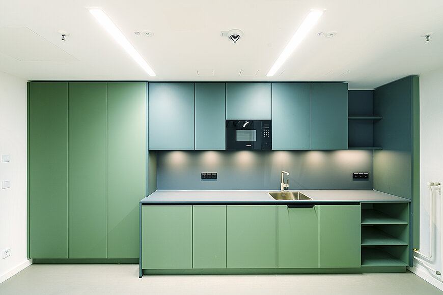 Nahaufnahme einer modernen Teeküche im sanierten Büro Flughafen Tempelhof. Die Küche hat glatte, mintgrüne Unterschränke und Hochschränke sowie petrolblaue Oberschränke mit integrierter Mikrowelle.