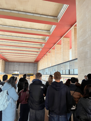 Schulklasse von hinten in der Haupthalle des Flughafen Tempelhof. Der Blickfang ist die markante Deckenkonstruktion aus hellen Holzbalken vor einem roten Hintergrund. Hohe Steinsäulen und eine verglaste Front auf der rechten Seite prägen den Raum, während im Hintergrund Anzeigetafeln zu erkennen sind.