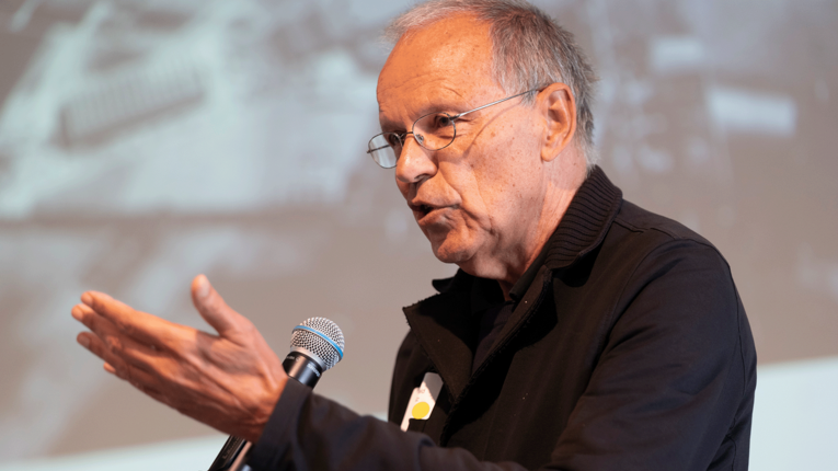 Dr. Harald Bodenschatz in einem schwarzen Pullover auf der Bühne bei THFxDenkmalwerkstatt. Er spricht in ein Mikrophon und streckt nad