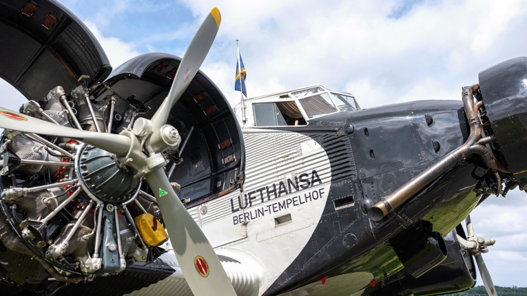 Nahaufnahme des historischen Flugzeugs Ju 52 auf dem "Lufthansa Berlin-Tempelhof" zu lesen ist. Die Maschine ist schwarz-weiß lackiert, im Hintergrund leicht bewölkter Himmel.