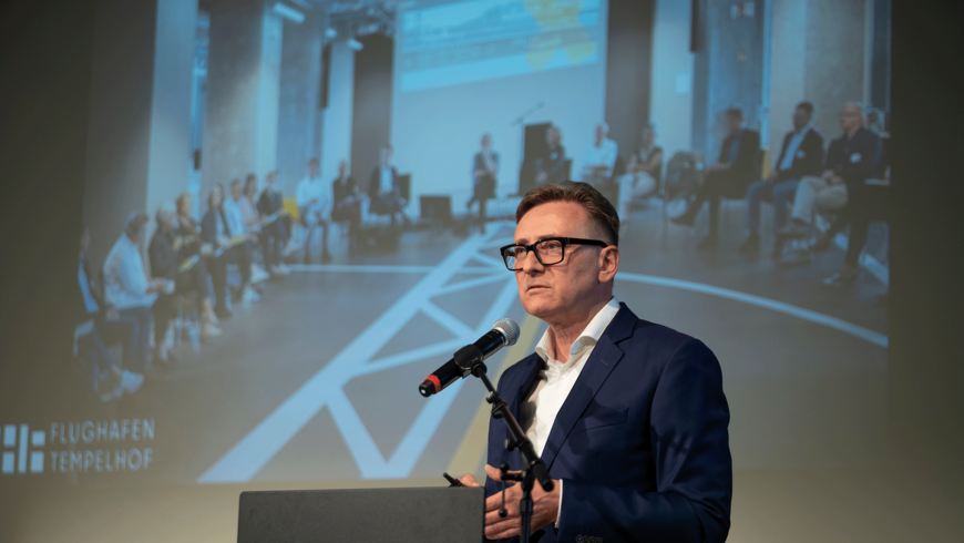 Ein Mann im blauen Anzug und mit Brille spricht in ein Mikrofon an einem dunklen Rednerpult. Im Hintergrund ist eine große Leinwand zu sehen, die eine Gruppe von Menschen zeigt, die in einer modernen, hellen Halle zusammensitzen. Unten links auf der Projektion ist ein Logo zu erkennen.