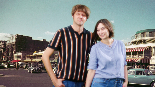 Die Band Grateful Cat steht vor einer Kulisse mit einer historischen Stadtstraße, alten Autos und Gebäuden mit Schildern und Markisen; sie tragen moderne Freizeitkleidung und bilden einen Kontrast zwischen Vergangenheit und Gegenwart.