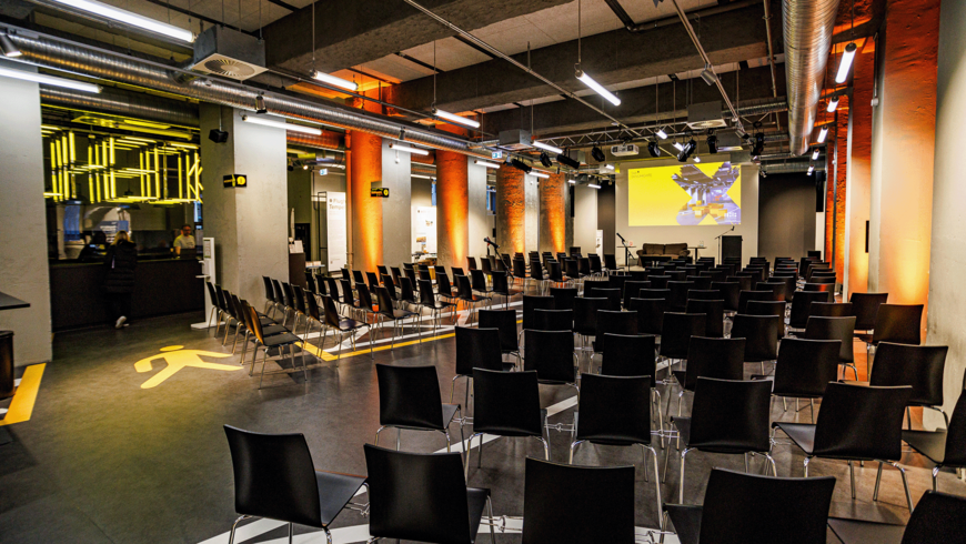 Blick in das Besucherzentrum CHECK-IN am Flughafen Tempelhof mit schwarzen Stühlen, Sichtbeton-Säulen und freiliegenden Lüftungsrohren. Im Hintergrund ist eine Leinwand mit gelbem Logo und eine Bühne zu sehen