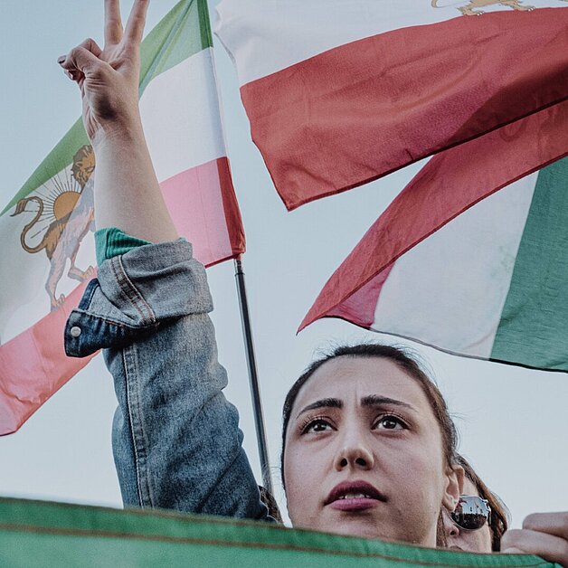 Person bei einer Demonstration hält zwei Finger zum Friedenszeichen erhoben. Im Hintergrund wehen mehrere Iran Flaggen.