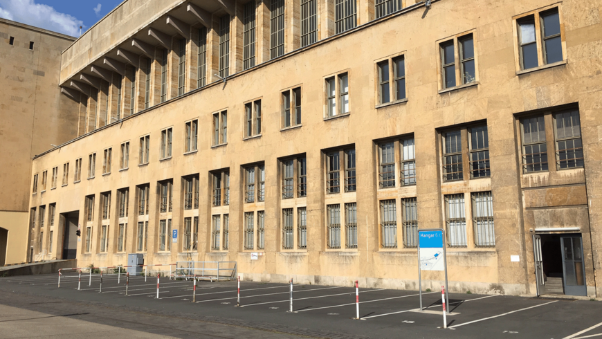 Außenansicht der langen, sandfarbenen Fassade des Flughafens Tempelhof. Die Wand erstreckt sich über zwei Stockwerke mit vielen, rechteckigen Fenstern, teils mit Gitter. Im Vordergrund befindet sich eine leere Reihe von Parkplätzen mit Absperrpfosten und einem blauen Schild, das auf den Hangar 6 hinweist.