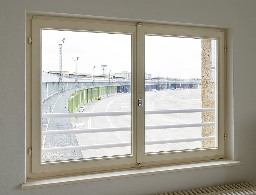 Blick aus einem Fenster des Büro Flughafen Tempelhof auf das Rollfeld bzw. Vorfeld, das durch eine grüne Brüstung und eine weiße Geländerkonstruktion begrenzt wird.