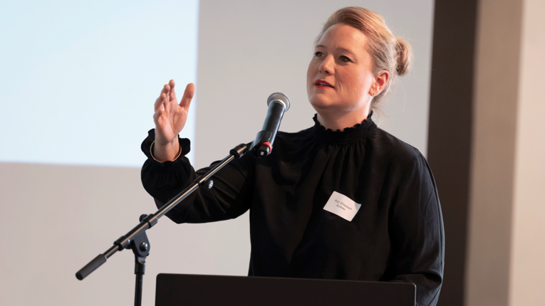 Prof. Dr. Elisabeth Endres steht hinter einem Podium und spricht in ein Mikrofon. Sie trägt dunkle Kleidung, hebt eine Hand zum Gestikulieren und präsentiert vor einer großen Projektionsfläche in einem Konferenz- oder Vortragsraum.