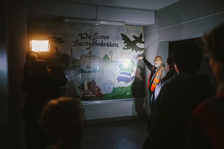 In einem dunkeln Raum steht eine Gruppe von Kinder und Erwachsenen vor einem Bild und leuchten es mit Taschenlampen an. Ein Guide mit roter Warnweste leuchtet auf das Bild