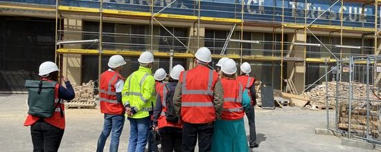 Besuchergruppe mit roten Warnwesten und weißen Bauhelmen steht vor einem Gebäude mit Baugerüst