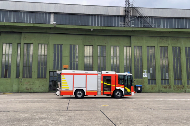 Rot-weißes Flughafenlöschfahrzeug steht auf einer großen Betonfläche vor einer historischen, grün gestrichenen Flugzeughalle mit hohen Fenstern am Flughafen Tempelhof.