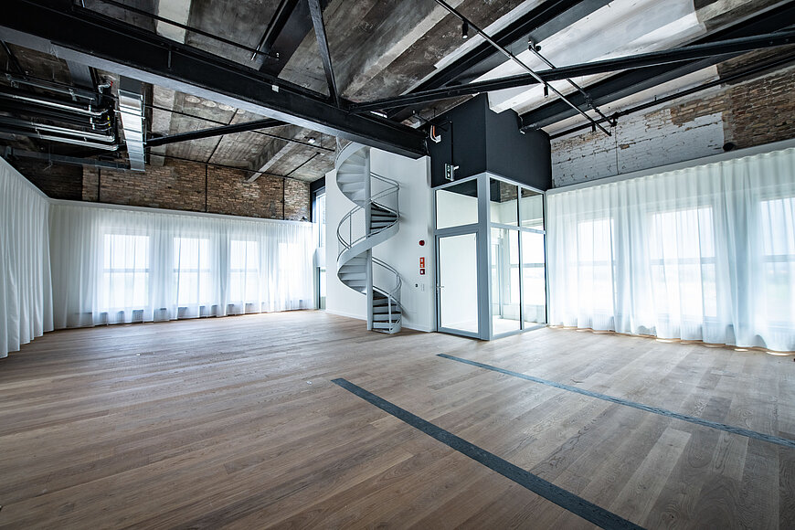 Die 6. Etage des THF TOWER mit Holzboden und hohen Decken. Die ursprüngliche Industriearchitektur ist sichtbar durch freiliegende Ziegelwände, dunkle Stahlträger und Lüftungsrohre unter der Decke. Eine elegante, weiße Wendeltreppe führt in eine obere Etage. Neben der Treppe befindet sich eine moderne, gläserne Aufzugskabine