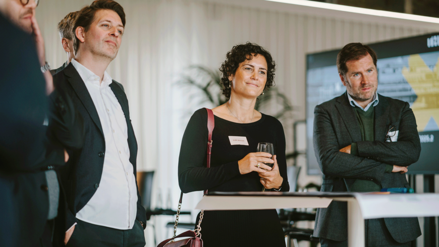 Mehrere Personen stehen bei der THFxDenkmalwerkstatt beieinander und blicken konzentriert nach vorne. Im Zentrum steht eine Frau mit dunklen Locken, die ein Weinglas hält. Die Atmosphäre wirkt professionell und interessiert.