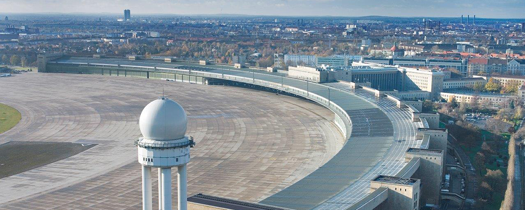 Luftaufnahme des Flughafen Tempelhof in Berlin mit dem halbkreisförmigen Terminalgebäude, den weitläufigen Vorfeldflächen und dem markanten weißen Radarturm im Vordergrund; im Hintergrund dicht bebaute Stadtbereiche unter leicht bewölktem Himmel.