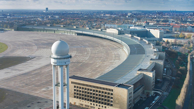 Luftaufnahme des Flughafen Tempelhof in Berlin mit dem halbkreisförmigen Terminalgebäude, den weitläufigen Vorfeldflächen und dem markanten weißen Radarturm im Vordergrund; im Hintergrund dicht bebaute Stadtbereiche unter leicht bewölktem Himmel.