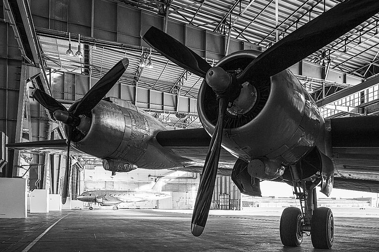 Schwarz-weiß Aufnahme der historischen Flugzeuge Iljuschin IL-14P, Douglas C-54 „Skymaster“ in einem der Hangar am Flughafen Tempelhof. Die große Hangartür ist geöffnet und gibt den Blick frei auf das Vorfeld und das Halbrund des Flughafengebäudes