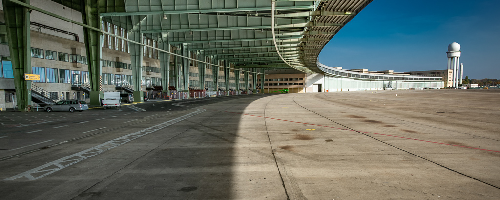 Vorfeld des Flughafen Tempelhof mit geschwungenem Dach und grünen Stahlträgern, davor eine breite Betonfläche im Schatten. Links stehen mehrere Fahrzeuge nahe der Fassade mit großen Fenstern. Im Hintergrund ragt ein weißer Radarturm mit kugelförmiger Kuppel in den klaren blauen Himmel