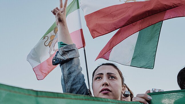 Person bei einer Demonstration hält zwei Finger zum Friedenszeichen erhoben. Im Hintergrund wehen mehrere Iran Flaggen.