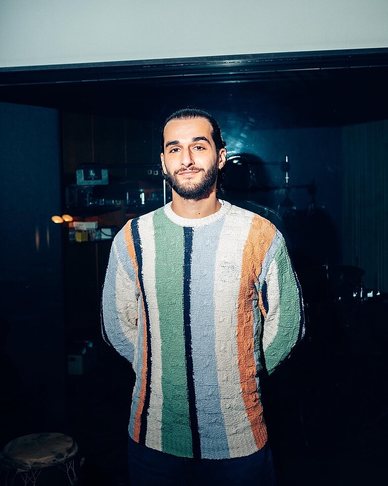 Marcel Moses steht in einem Innenraum vor einer dunklen Glaswand, trägt einen mehrfarbig gestreiften Pullover und ist klar ausgeleuchtet, während hinter der Scheibe Musikinstrumente wie ein Schlagzeug schemenhaft erkennbar sind.