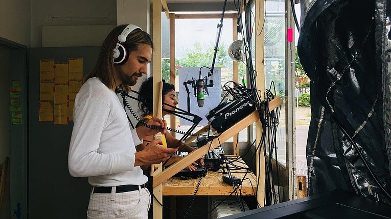 Im Torhaus am Flughafen Tempelhof steht ein Mann mit Kopfhörern vor einem Mischpult, dahinter sitzt eine Frau vor einem Mikrophon