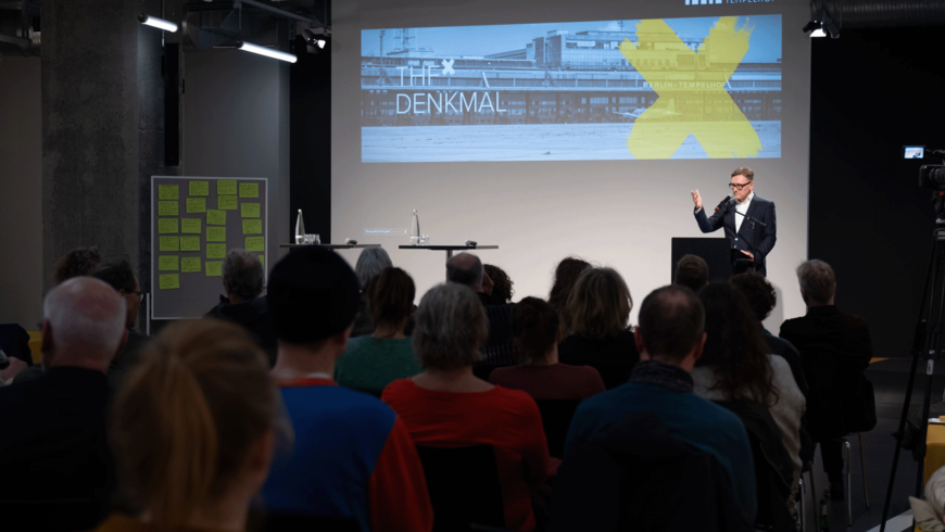 Eine Person steht an einem Podium und präsentiert vor einem sitzenden Publikum. Hinter ihr ist eine große Leinwand mit einer Folie zu sehen, die ein Brücken- oder Stahlkonstruktionsmotiv mit einem gelben X zeigt. Der Raum wirkt modern und professionell, mit Beleuchtung, Bannern und einem Betonpfeiler an der Seite.