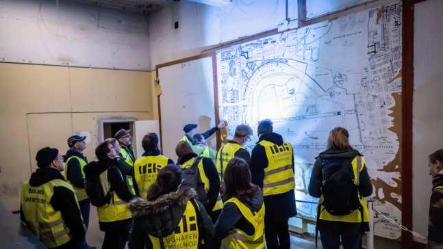 Mehrere Personen in gelben Warnwesten mit den Aufdrucken "Flughafen Tempelhof“ und „Berliner Unterwelten“ stehen vor einer großen Wandkarte des Flughafen Tempelhof. Eine Person zeigt auf einen Bereich des Plans, während die Gruppe aufmerksam zuhört. Die Szene wirkt wie eine geführte Erläuterung zur historischen Anlage.