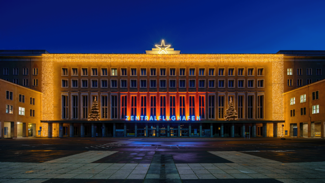 Weihnachtlich beleuchtete Fassade des Flughafen Tempelhof bei Nacht mit farbigen Lichtakzenten vor dem zentralen Eingangsbereich und leerem Vorplatz.