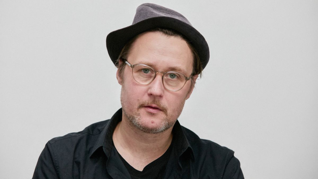 Porträt des Künstlers Mittekill vor hellem Hintergrund. Er trägt eine dunkle Brille, ein dunkelgraues Hutmodell und ein dunkles Oberteil. Der Blick ist ruhig und direkt in die Kamera gerichtet.
