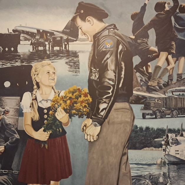 Gemälde am Flughafen Tempelhof mit mehreren Szenen der Luftfahrt und humanitärer Hilfe: Im Vordergrund lächelt ein Pilot ein Mädchen mit Blumen an; im Hintergrund verschiedene Flugzeuge, Fahrzeuge und Menschen beim Verladen von Hilfsgütern sowie Kinder, die den ankommenden Maschinen zuwinken.