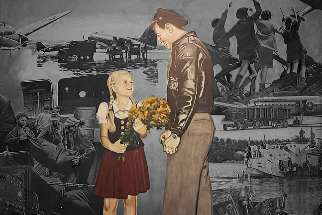 Foto von dem Gemälde in der Haupthalle des Flughafen Tempelhof, auf dem ein Soldat zu einem Mädchen runterschaut, das einen Blumenstrauß in der Hand hält und barfuß ist
