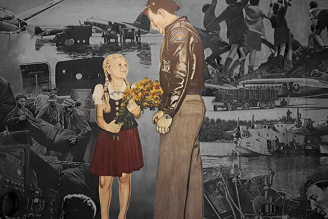 Foto von dem Gemälde in der Haupthalle des Flughafen Tempelhof, auf dem ein Soldat zu einem Mädchen runterschaut, das einen Blumenstrauß in der Hand hält und barfuß ist