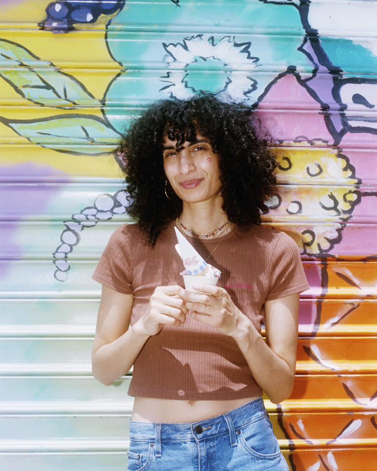 Sarah Farina steht vor einem farbenfrohen Blumen‑Wandbild, trägt ein braunes kurzes Oberteil und Jeans und hält einen Becher Eis mit Löffel und Waffel in der Hand.