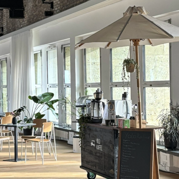 Mobile Kaffeebar mit einem Schrim im THF TOWER. Im Hintergrund Tische und Stühle sowie grüne Pflanzen. Durch die großen Fenster fällt Licht in das Café