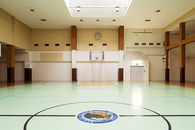 Blick in die leere Basketballhalle am Flughafen Tempelhof. Im Hintergrund befindet sich ein Korb, darüber eine Uhr. Durch ein fenster in Decke kommt Licht hinein