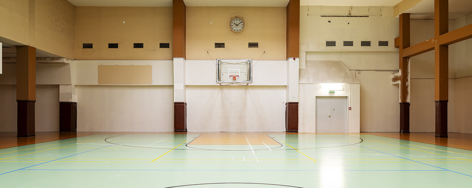 Blick in die leere Basketballhalle am Flughafen Tempelhof. Im Hintergrund befindet sich ein Korb, darüber eine Uhr. Durch ein fenster in Decke kommt Licht hinein