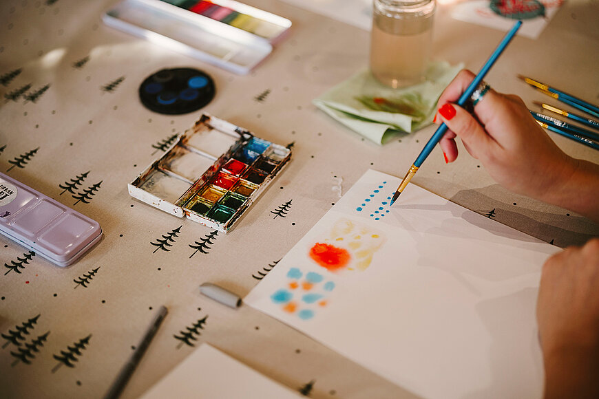 Bild von einer Hand mit einem Pinsel. Es werden Aquarelle auf einem weißen Blatt gemalt, daneben ein Tuschkasten