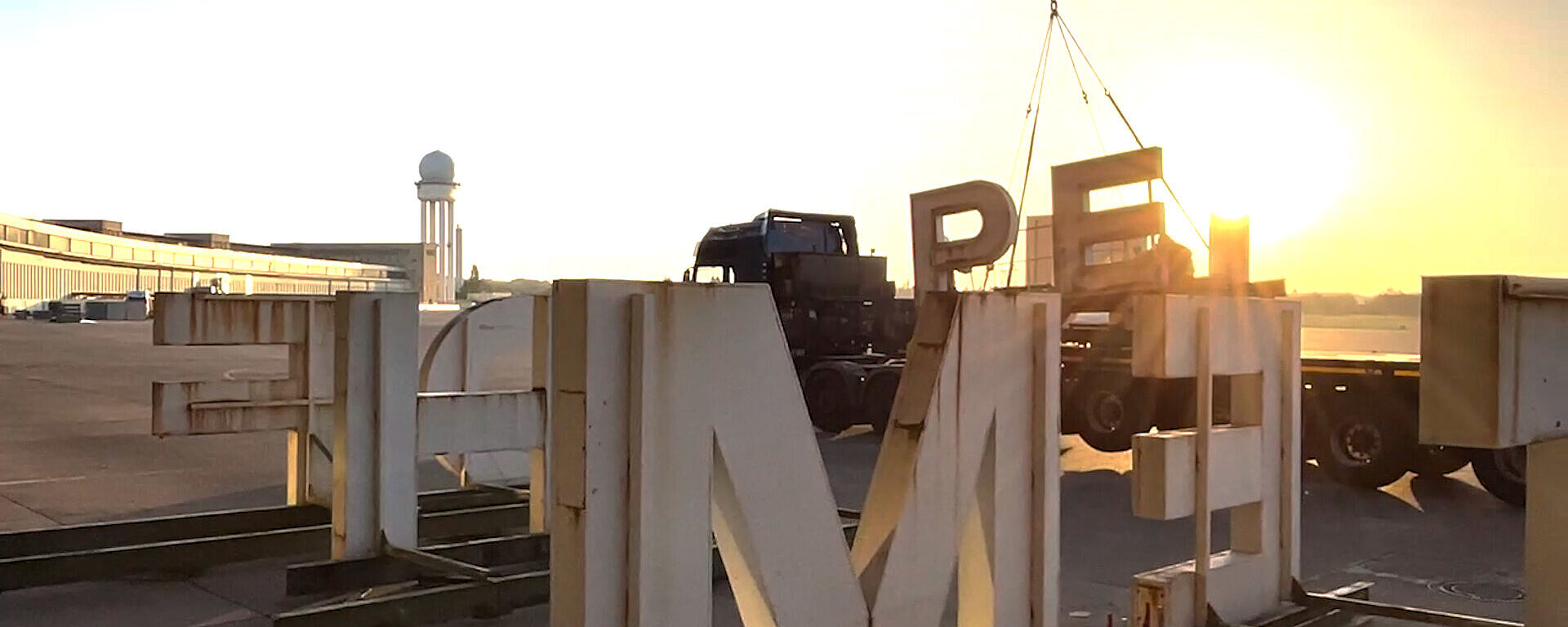 Der Schriftzug Berlin Tempelhof wird mit einem Kran abgebaut. Im Hintergrund geht die Sonne auf