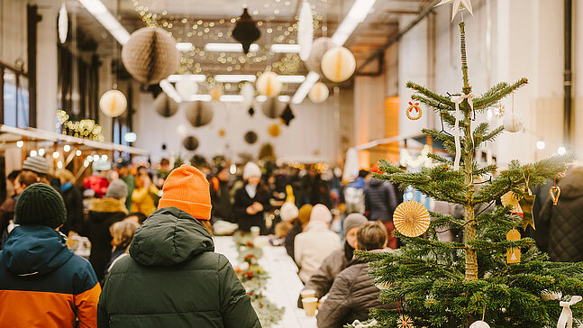 Blick in die winterlich geschmückte Alte Feuerwache am Flughafen Tempelhof. Im Vordergrund steht ein geschmückter Tannenbaum und im Hintergrund sieht man viele Menschen mit dicken Jacken und Mützen